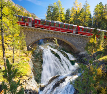 Rødt schweizisk tog kører over stenbro med vandfald i Zermatt under efterårsfarver
