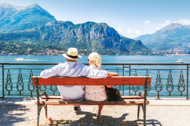 Ældre par nyder udsigten over Comosøen fra en træbænk med bjerglandskab og roligt vand i Norditalien
