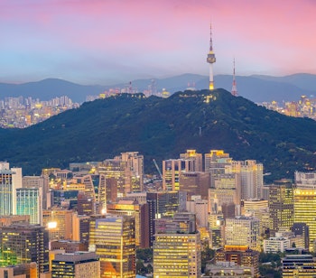 Imponerende udsigt over Seouls bymidte ved solnedgang med oplyste skyskrabere mod bjergkulissen, der viser Sydkoreas moderne hovedstad og økonomiske udvikling