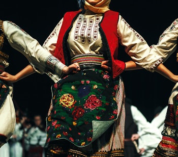 Tre kvinder i farverige serbiske folkedragter med broderede forklæder danser traditionel folkedans fra Balkan