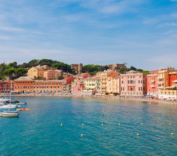Den idylliske Baia del Silenzio i Sestri Levante, Ligurien, med farverige italienske bygninger langs strandpromenaden, krystalklart turkisblåt vand og små fiskerbåde ved den gyldne strand