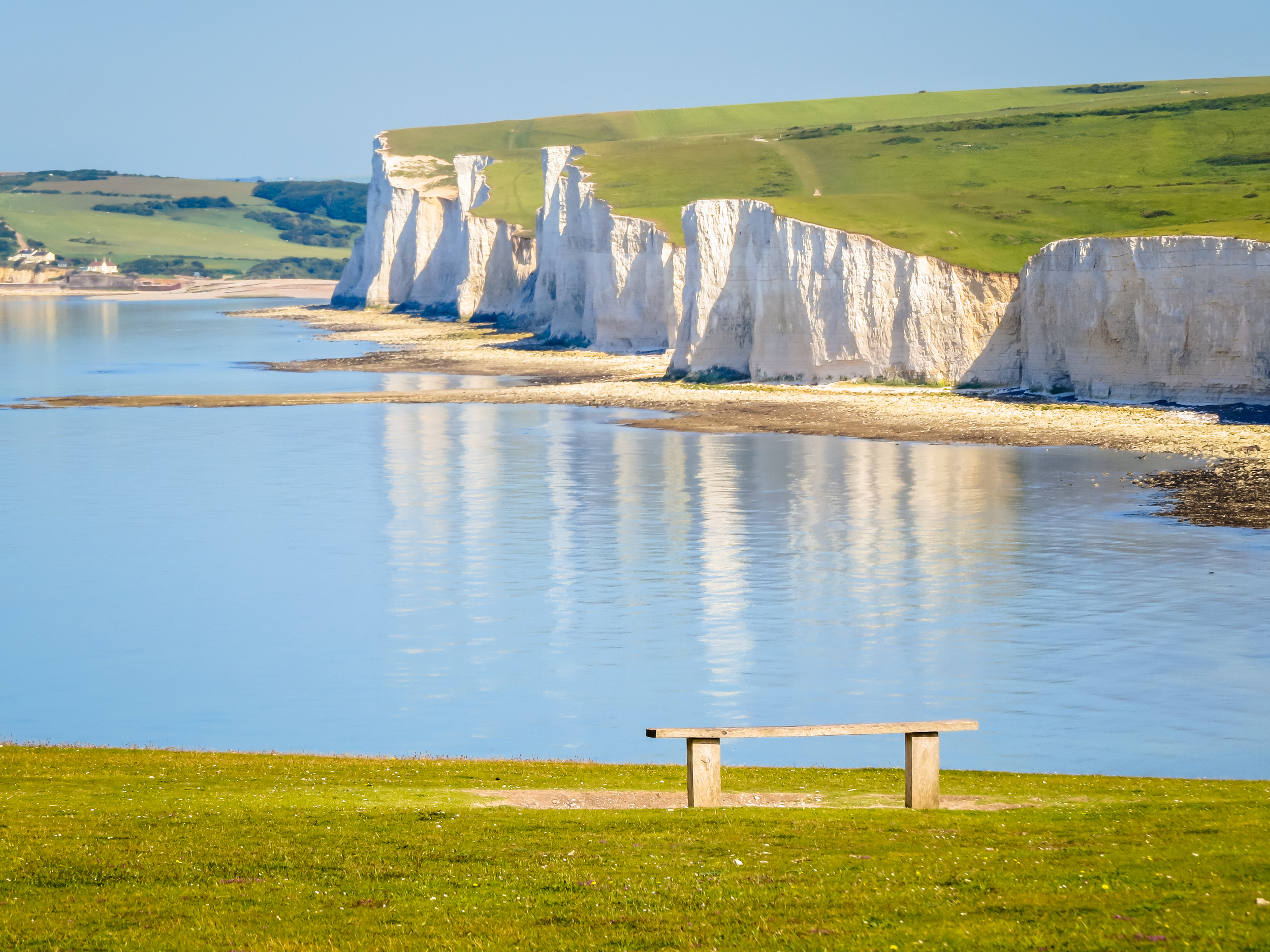 De ikoniske Seven Sisters kridtklipper langs kysten i East Sussex, England, spejlet i det rolige blå hav på en solrig sommerdag med en træbænk i forgrunden