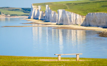 De ikoniske Seven Sisters kridtklipper langs kysten i East Sussex, England, spejlet i det rolige blå hav på en solrig sommerdag med en træbænk i forgrunden