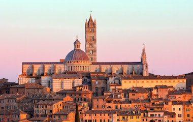 Den majestætiske Siena Katedral med dens karakteristiske stribede marmorfacade badet i gyldent solnedgangslys over de terracottafarvede tage i den historiske toscanske by