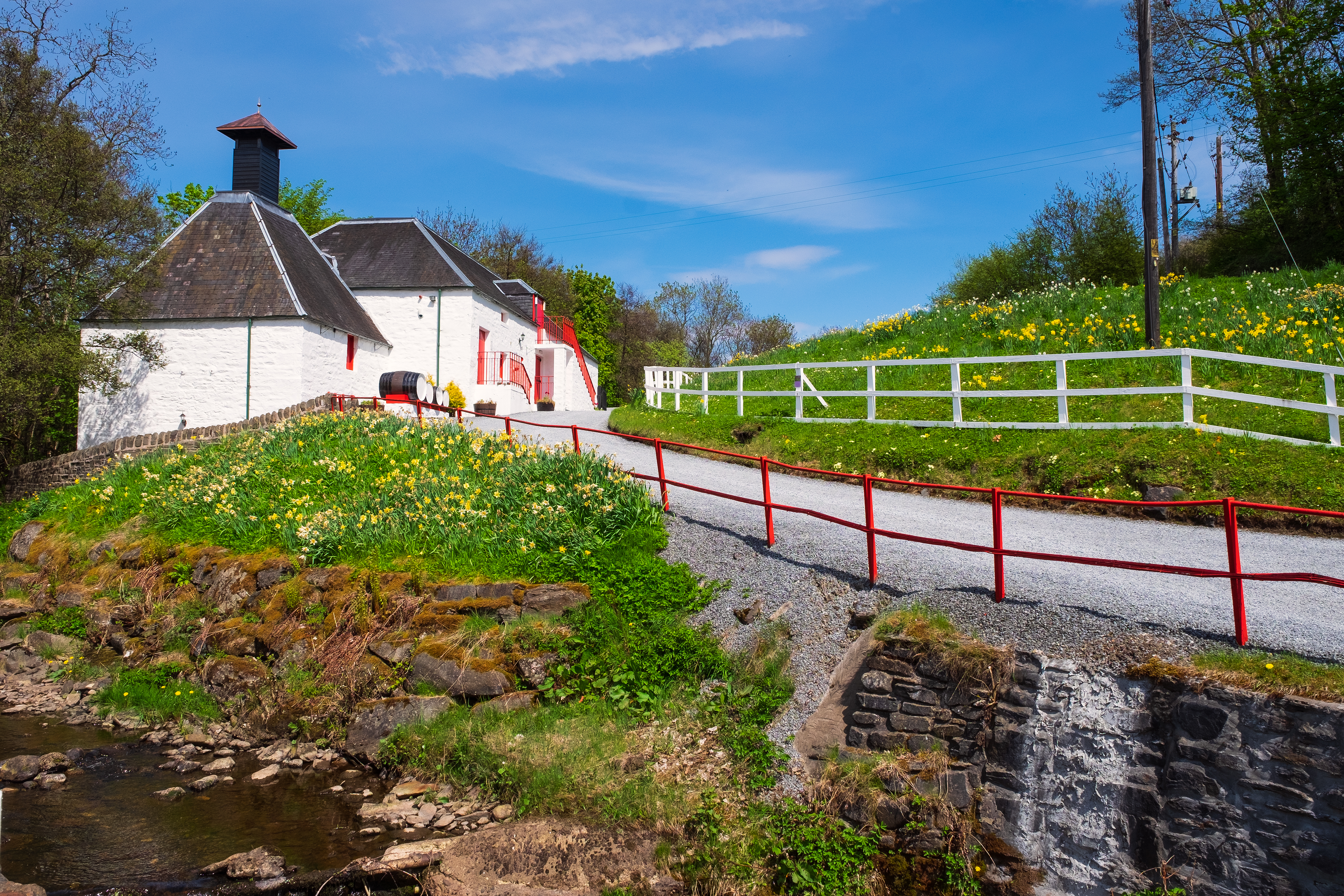 Idyllisk hvidt whisky-destilleri i de skotske Highlands nær Pitlochry med traditionelt pagodetag, rød hegning og blomstrende grønne bakker langs en lille bæk på en smuk sommerdag