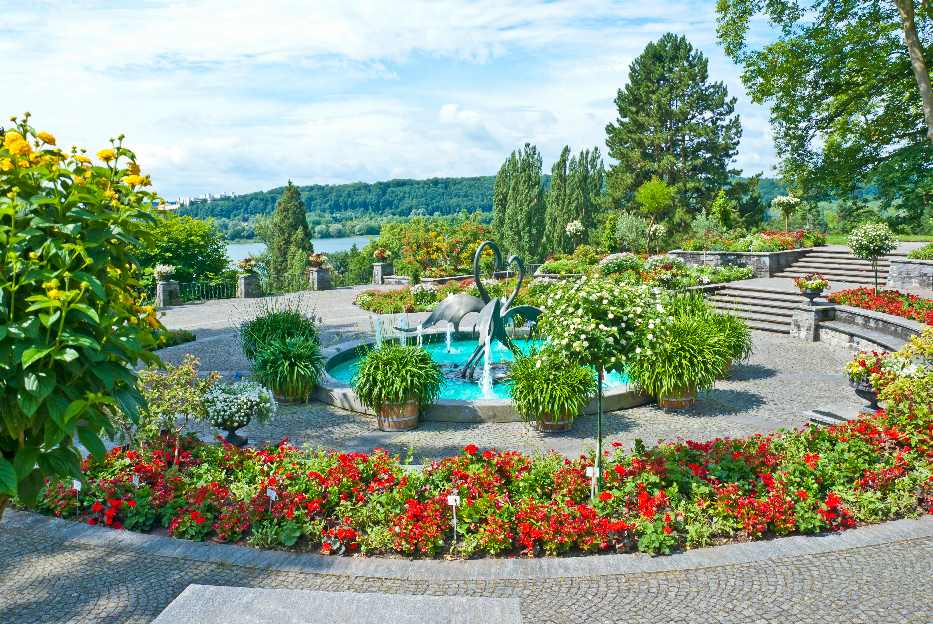 Smuk søterrasse med haveskulptur og farverige blomsterbede med panoramaudsigt over sø og bjerge