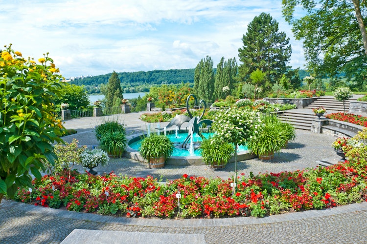 Smuk søterrasse med haveskulptur og farverige blomsterbede med panoramaudsigt over sø og bjerge