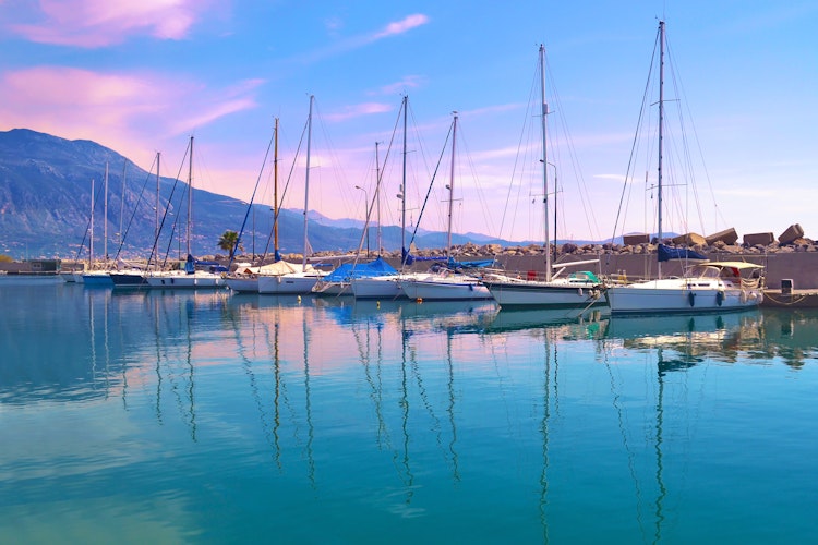 Magisk solnedgang over Kalamata havn på Peloponnes med farverige sejlbåde, der spejler sig i det stille, turkisblå vand under en lilla aftenhimmel