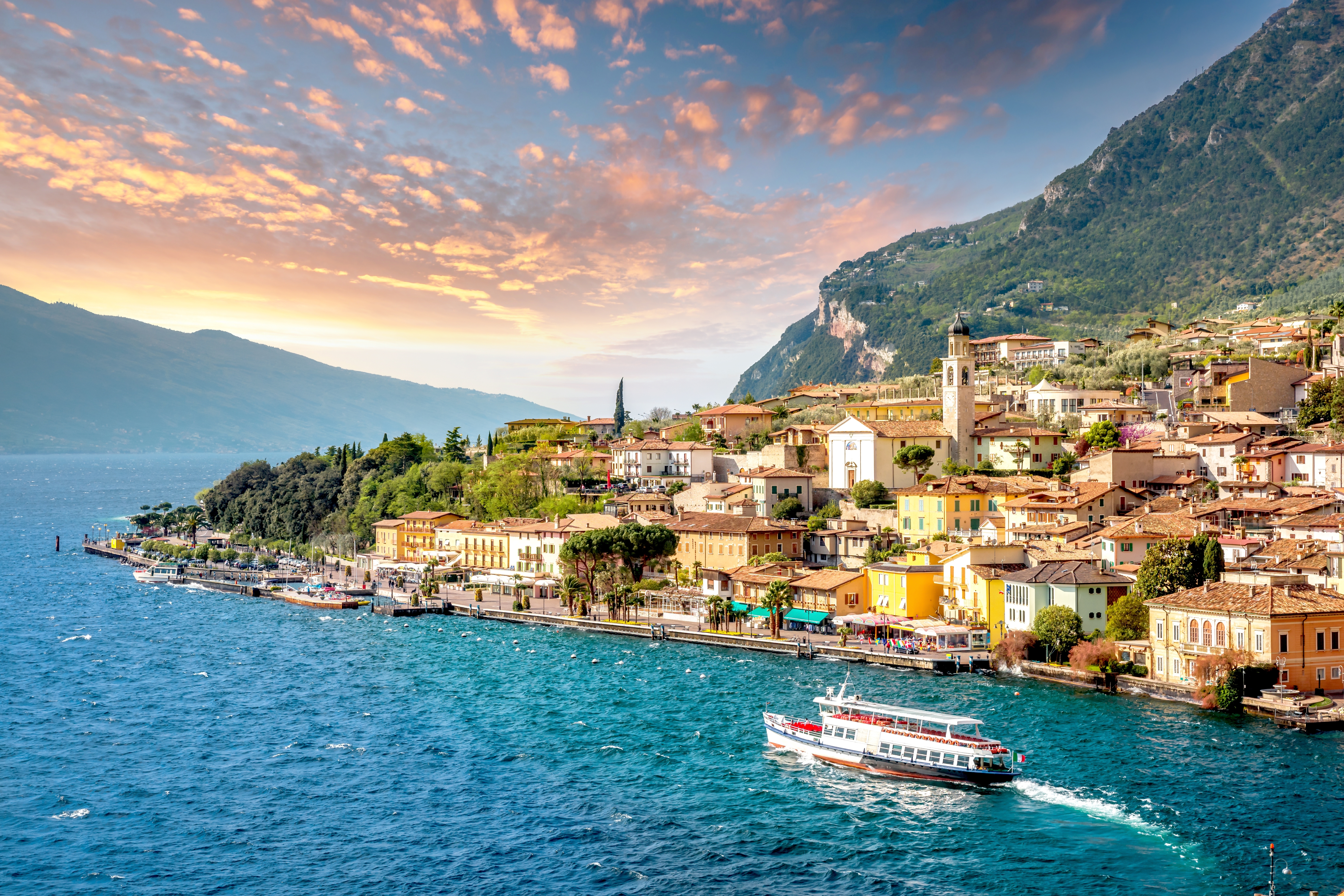 Idyllisk panoramaudsigt over Limone sul Garda med farverige bygninger ved Gardasøens blå vand og majestætiske bjerge i solnedgangslys