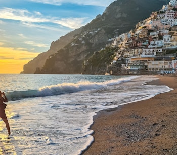 Kvinde i silhuet ved solnedgang på Marina Grande stranden med Positanos farverige bygninger på skrænten ved Amalfikysten i Italien.