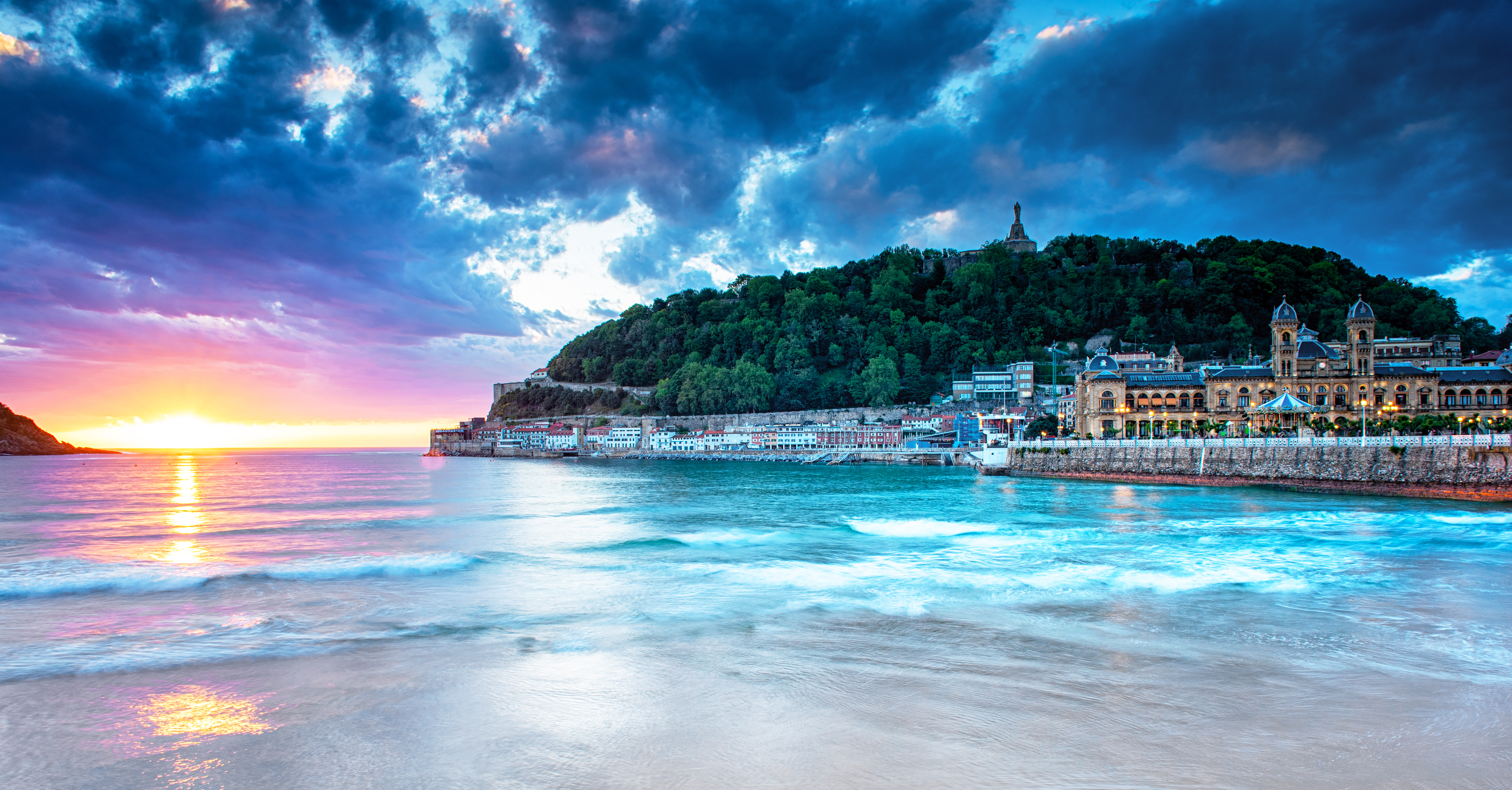 Smuk solnedgang over stranden i San Sebastian, Spanien, med turkisblåt vand og historiske bygninger langs kysten