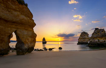 Betagende solopgang over den berømte Camilo-strand i Algarve, Portugal, med gyldne klipper, naturlige stenbuer og krystalklart turkisblåt vand