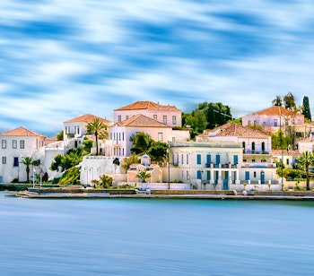 Idyllisk kystlinje på Spetses-øen i den Saroniske Bugt, der viser traditionelle hvide bygninger med terrakottatage langs det krystalklare blå vand, perfekt til en rolig græsk ferie