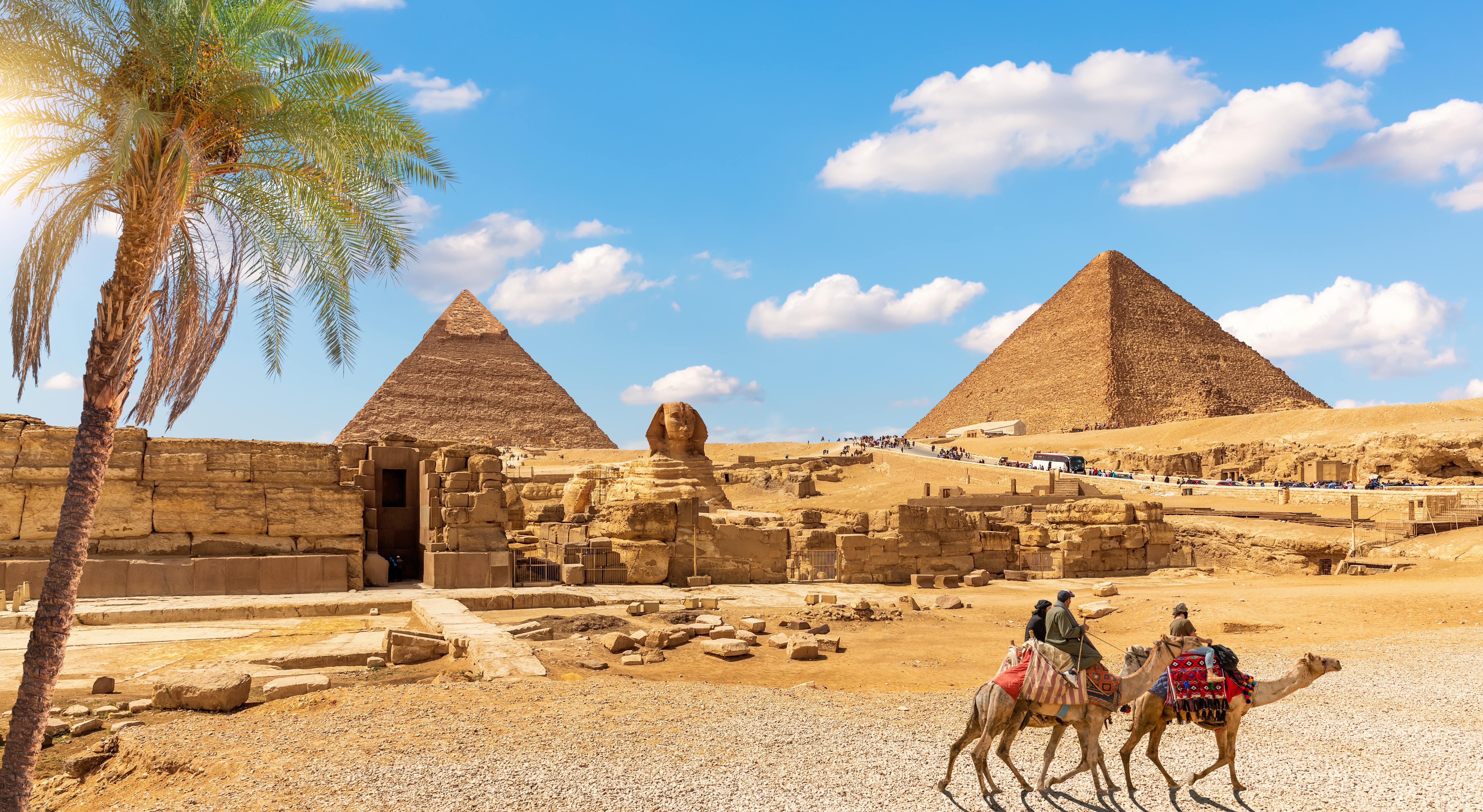 Den store Sphinx og pyramiderne i Giza med kamelryttere i forgrunden og en palme under blå himmel, Egypten