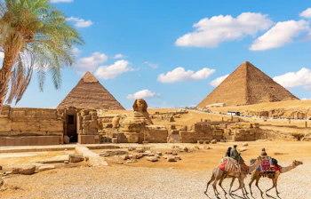 Den store Sphinx og pyramiderne i Giza med kamelryttere i forgrunden og en palme under blå himmel, Egypten
