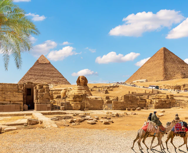 Den store Sphinx og pyramiderne i Giza med kamelryttere i forgrunden og en palme under blå himmel, Egypten