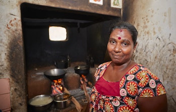 Srilankansk kvinde i traditionel beklædning tilbereder mad over et brændefyret komfur i et simpelt landsbykøkken, der viser autentisk landlig livsstil