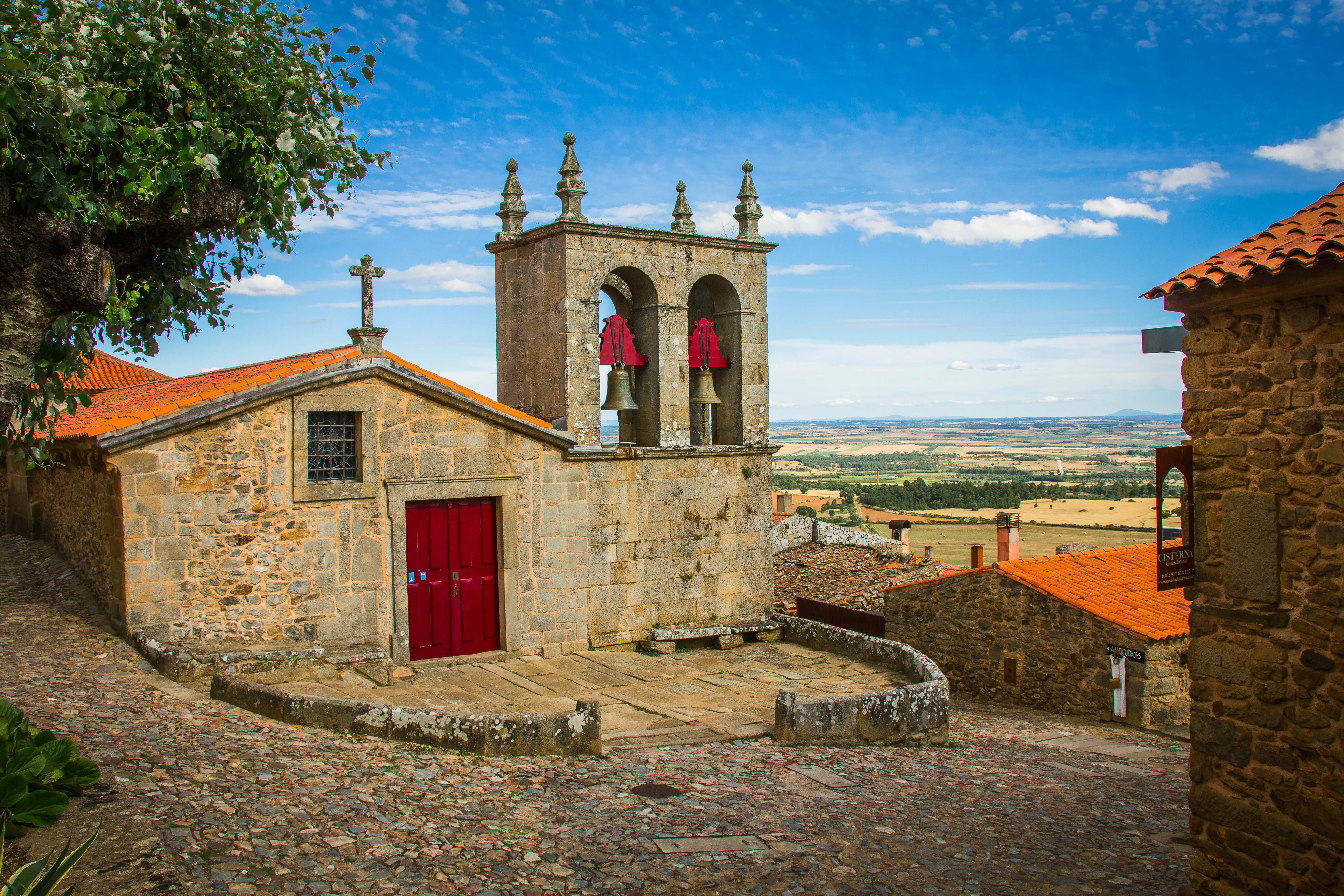 Gammel stenkirke med rødt klokketårn og brolagt plads i Figueira de Castelo Rodrigo, Portugal, med udsigt over grønt landskab