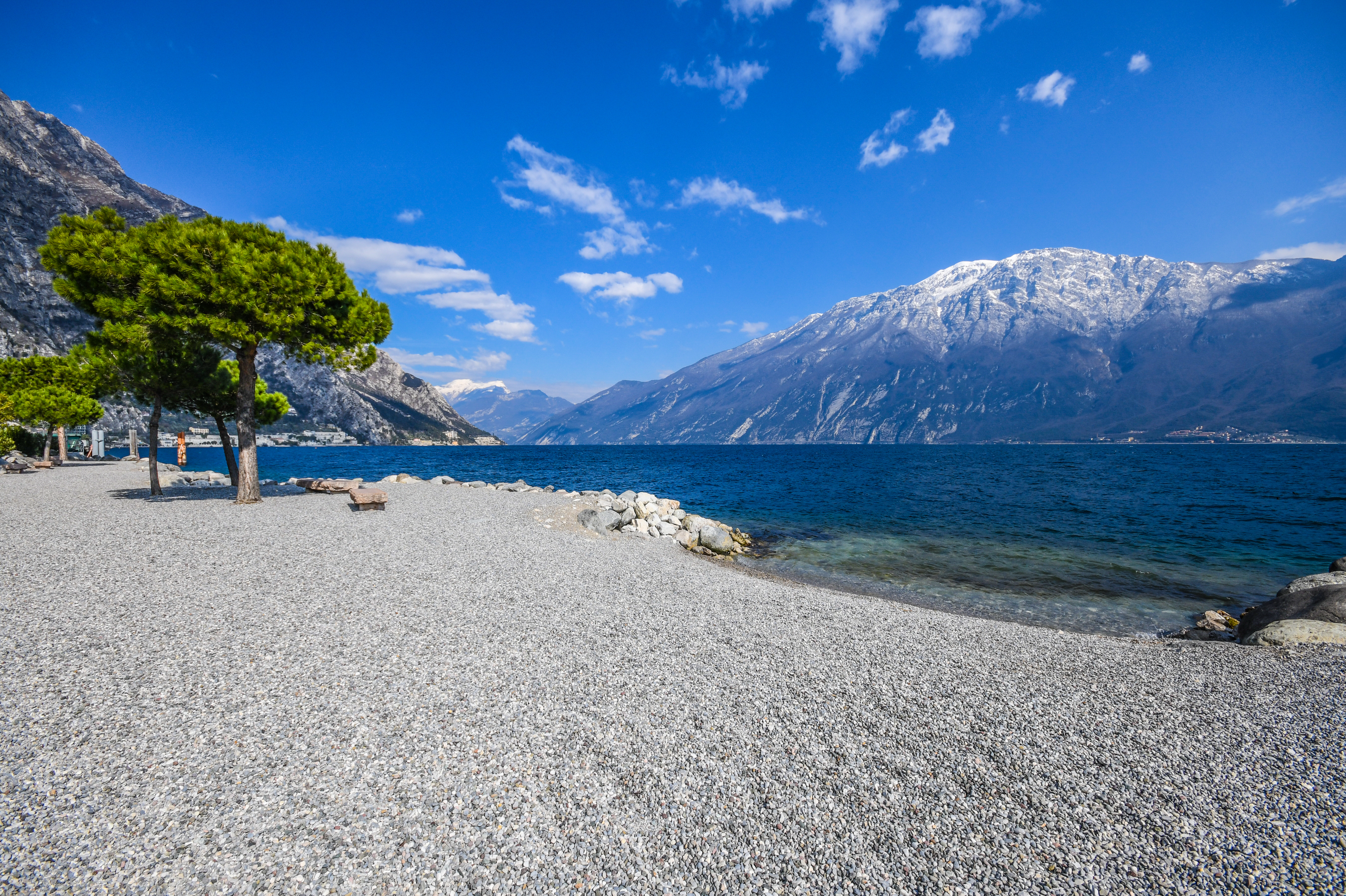 Idyllisk stenstrand ved Gardasøen med krystalklart vand og majestætiske bjerge i baggrunden. Perfekt til en afslappende dag i naturskønne omgivelser