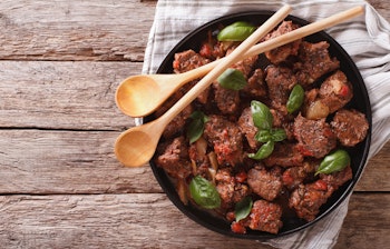 Traditionel græsk Stifado med mørt oksekød i en aromatisk tomatsauce garneret med frisk basilikum, serveret i en rustik skål på et træbord