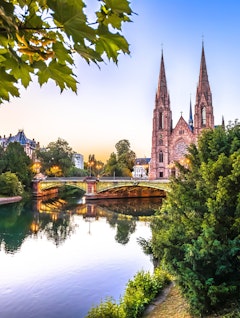 Idyllisk solnedgangsudsigt over Saint Paul kirken i Strasbourg med dens tvillingtårne, der spejler sig i kanalen omgivet af grønne træer og en charmerende stenbro i Alsace-regionen