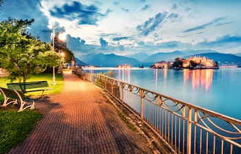 Stemningsfuld aftenudsigt over Stresa med den oplyste strandpromenade langs Lago Maggiore ved solnedgang, og den berømte Isola Bella ø i baggrunden