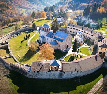 Betagende luftfoto af det historiske Studenica-kloster i Serbien badet i gyldent solnedgangslys, med den hvide marmorkirke omgivet af farverige efterårstræer i det balkanske bjerglandskab