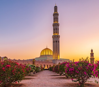 Den storslåede Sultan Qaboos Grand Mosque i Muscat, Oman ved solnedgang med gyldent lys over kupler, minareter og de smukt anlagte haver