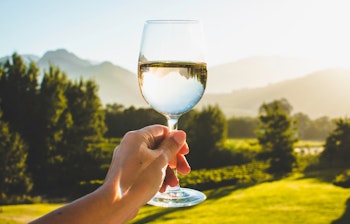 Hånd der holder et glas hvidvin mod den smukke solnedgang over vinmarkerne i Franschhoek, Sydafrika