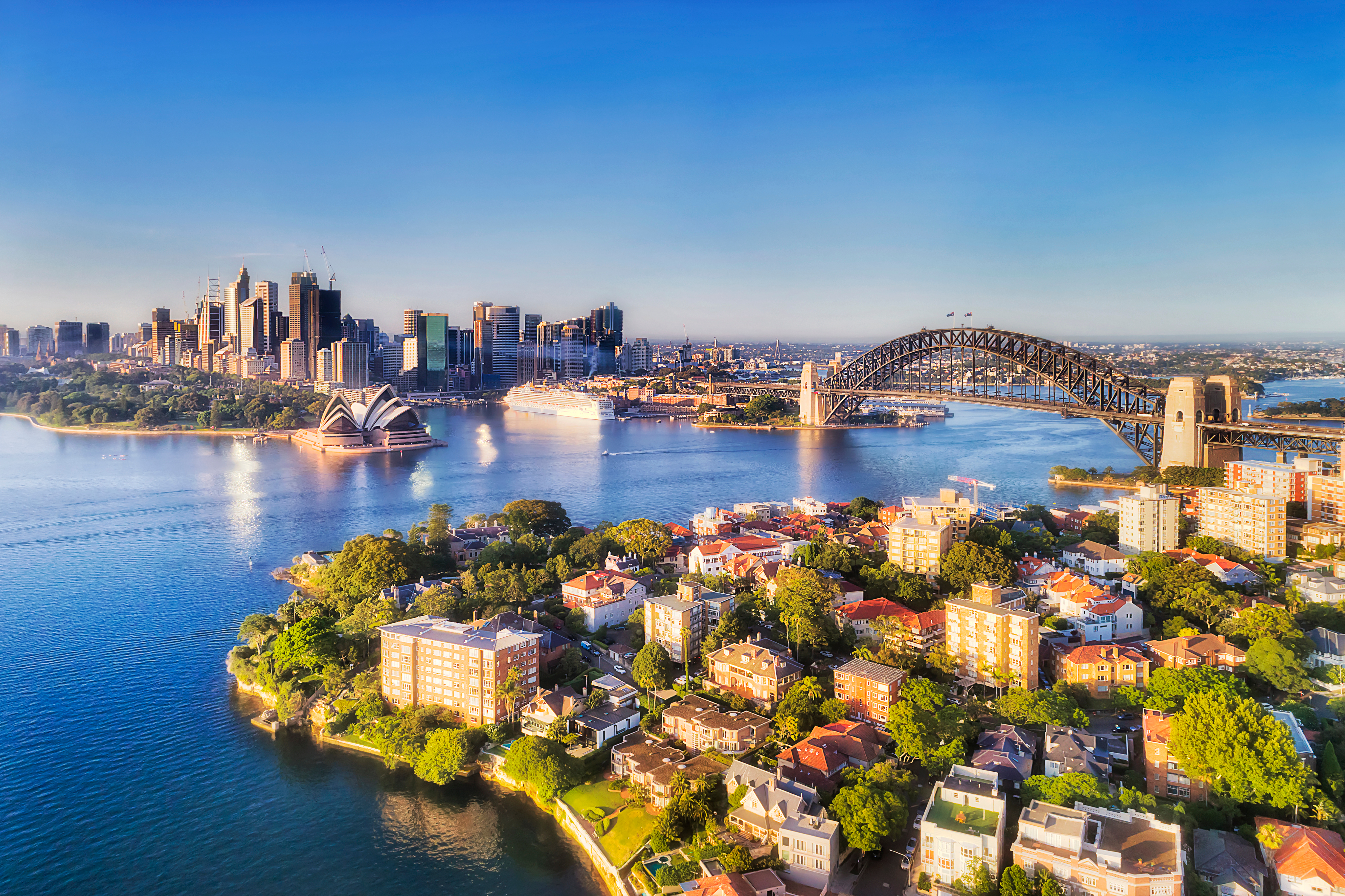 Betagende luftfoto af Sydney Harbour på en solrig dag, med den ikoniske Sydney Harbour Bridge, der forbinder CBD'ens skyskrabere med North Shore-forstæderne, omgivet af blåt havnevand og eksklusive ejendomme ved vandkanten
