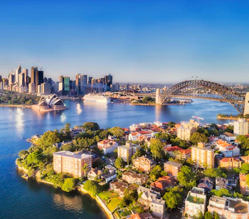 Betagende luftfoto af Sydney Harbour på en solrig dag, med den ikoniske Sydney Harbour Bridge, der forbinder CBD'ens skyskrabere med North Shore-forstæderne, omgivet af blåt havnevand og eksklusive ejendomme ved vandkanten