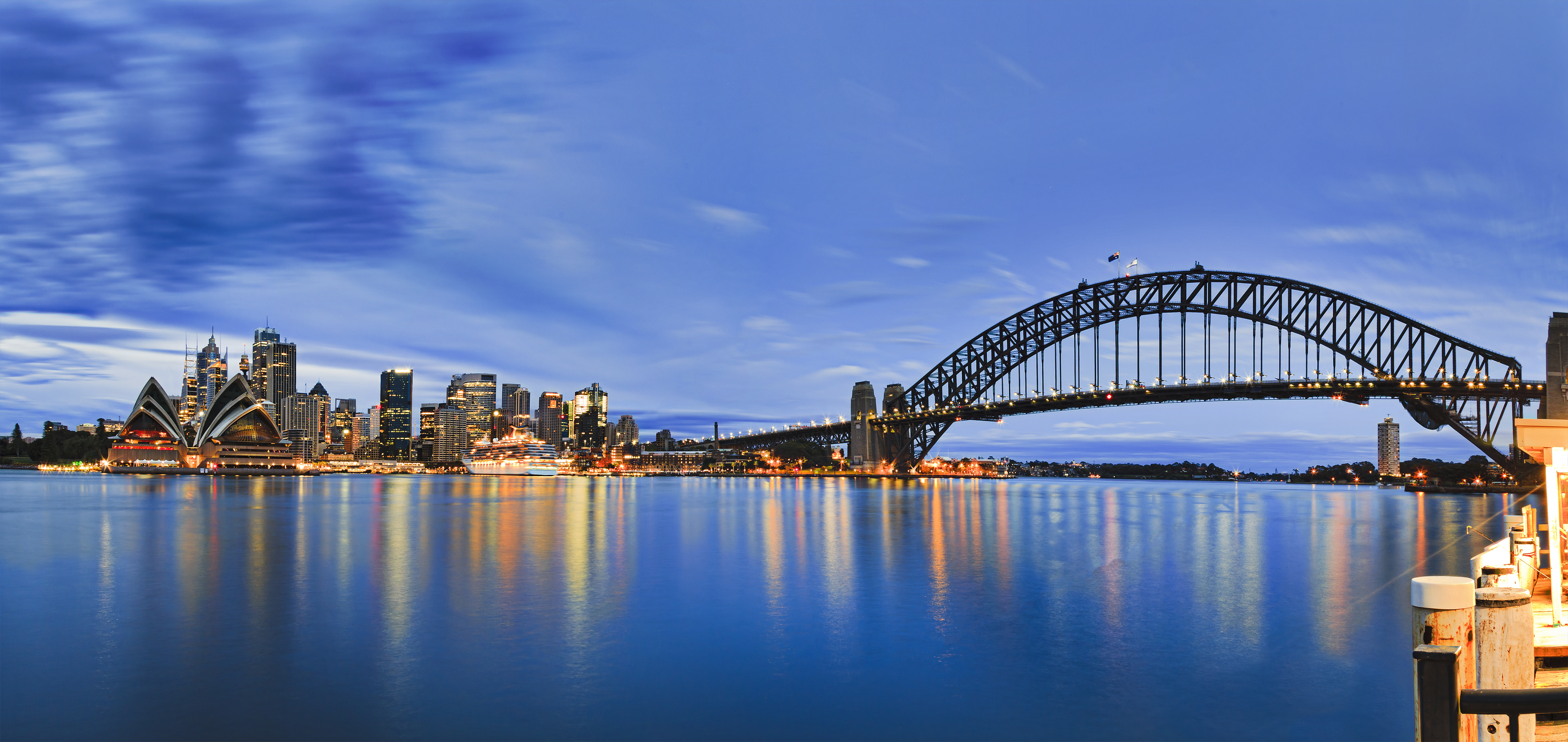 Betagende panoramaudsigt over Sydneys skyline ved solopgang med den ikoniske Harbour Bridge og Operahuset, der spejler sig i havnens stille vand. Byens højhuse og Kirribilli-molen ses i det blå morgenlys.