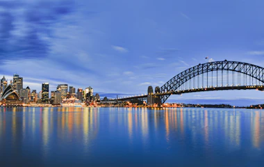Betagende panoramaudsigt over Sydneys skyline ved solopgang med den ikoniske Harbour Bridge og Operahuset, der spejler sig i havnens stille vand. Byens højhuse og Kirribilli-molen ses i det blå morgenlys.