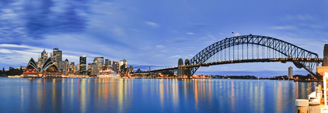 Betagende panoramaudsigt over Sydneys skyline ved solopgang med den ikoniske Harbour Bridge og Operahuset, der spejler sig i havnens stille vand. Byens højhuse og Kirribilli-molen ses i det blå morgenlys.