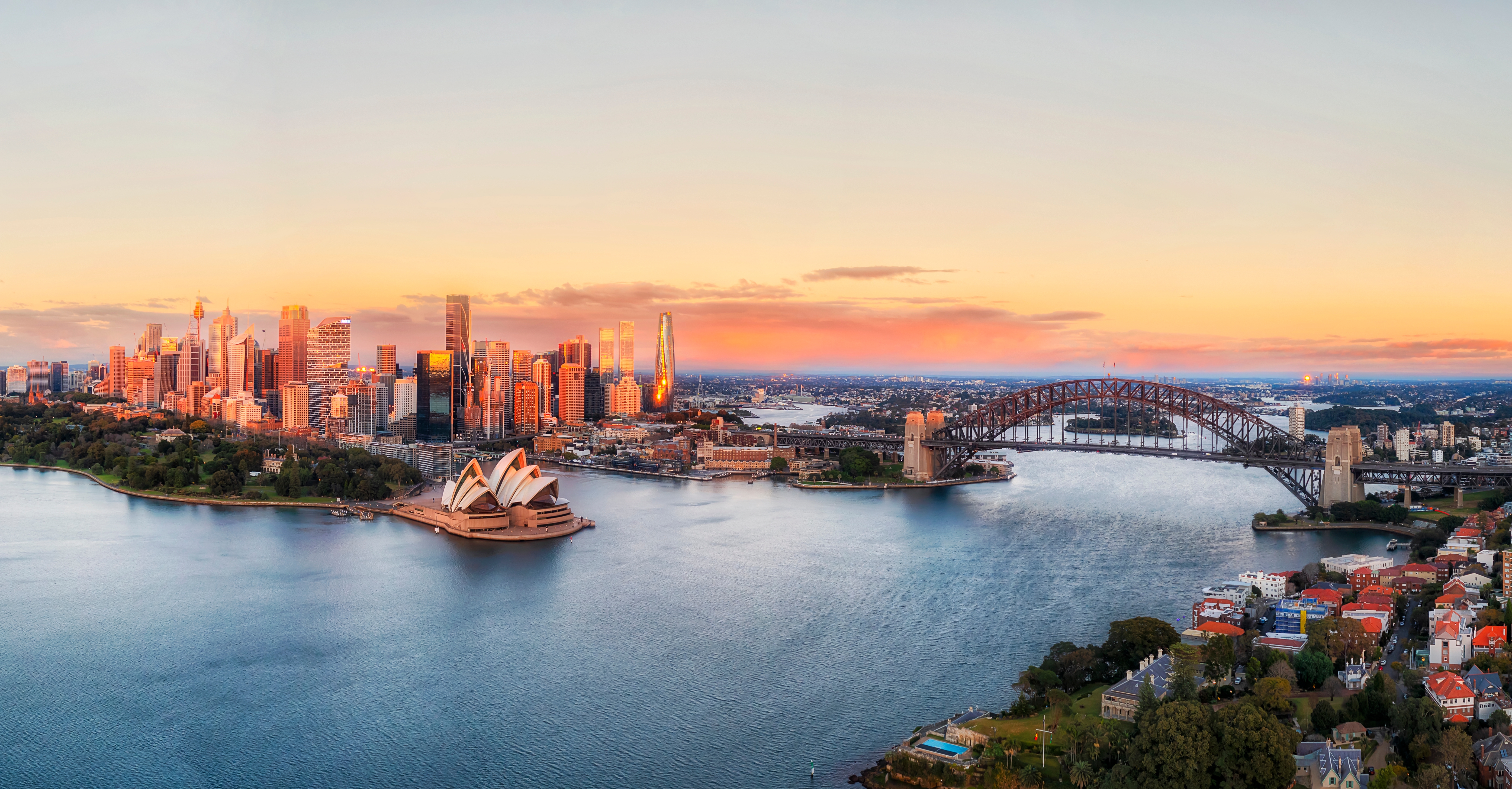 Betagende luftfoto af Sydney i solopgang med det ikoniske Operahus, Harbour Bridge og byens skyline, hvor det gyldne morgenlys reflekteres i havnens vand
