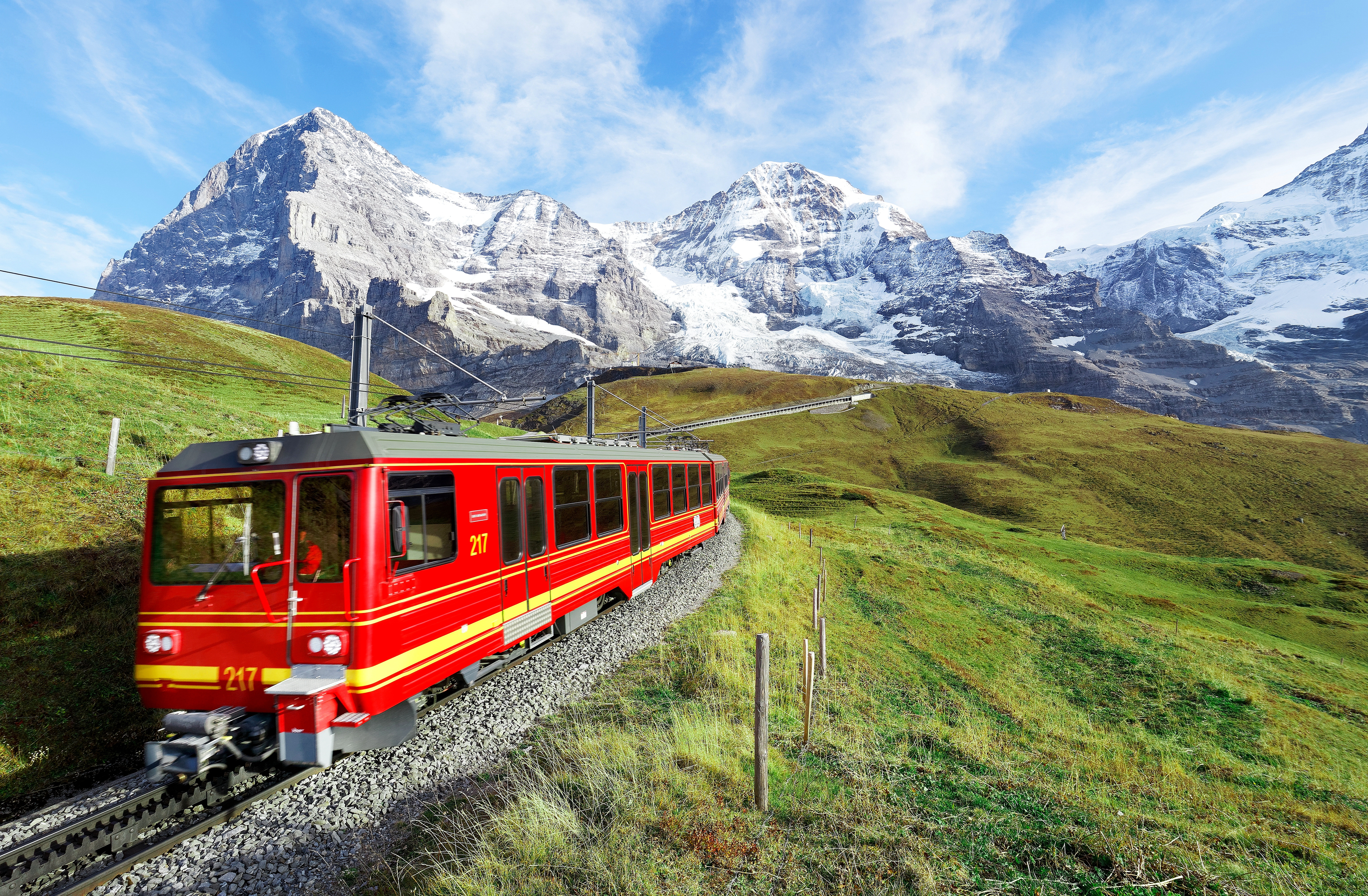 Rødt tandhjulstog på rejse gennem grønne alpeenge fra Jungfraujoch mod Kleine Scheidegg med Eiger, Mönch og Jungfrau bjergene i baggrunden, Berner Oberland, Schweiz
