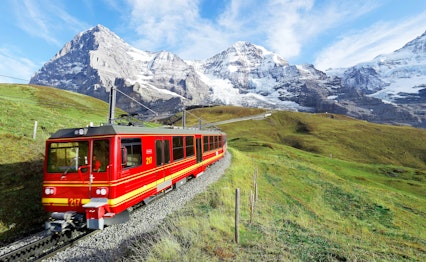 Rødt tandhjulstog på rejse gennem grønne alpeenge fra Jungfraujoch mod Kleine Scheidegg med Eiger, Mönch og Jungfrau bjergene i baggrunden, Berner Oberland, Schweiz