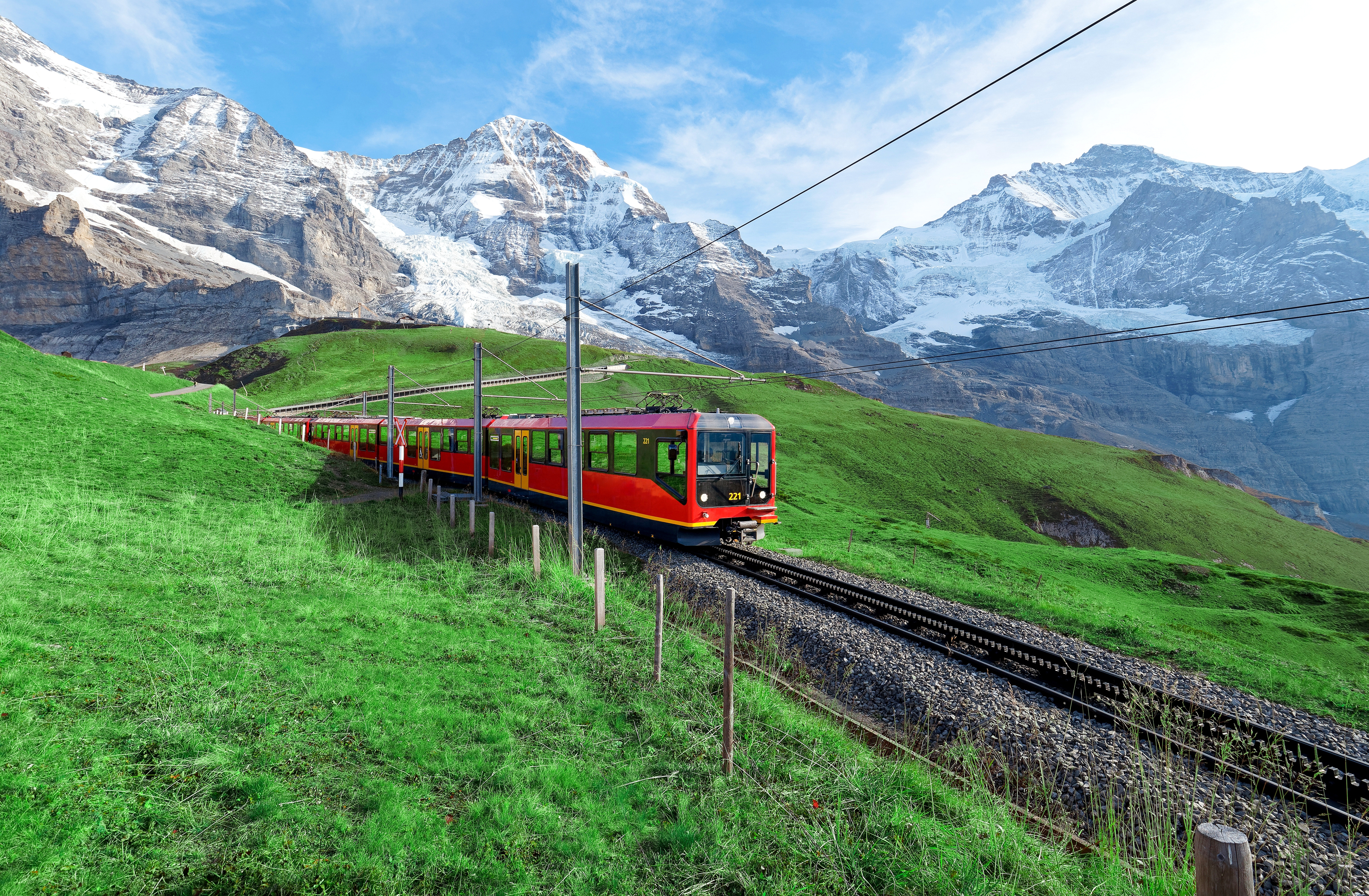Rødt tandhjulstog på vej fra Jungfraujoch til Kleine Scheidegg gennem grønne alpinenge med de majestætiske Jungfrau og Mönch bjerge i baggrunden i Schweiz