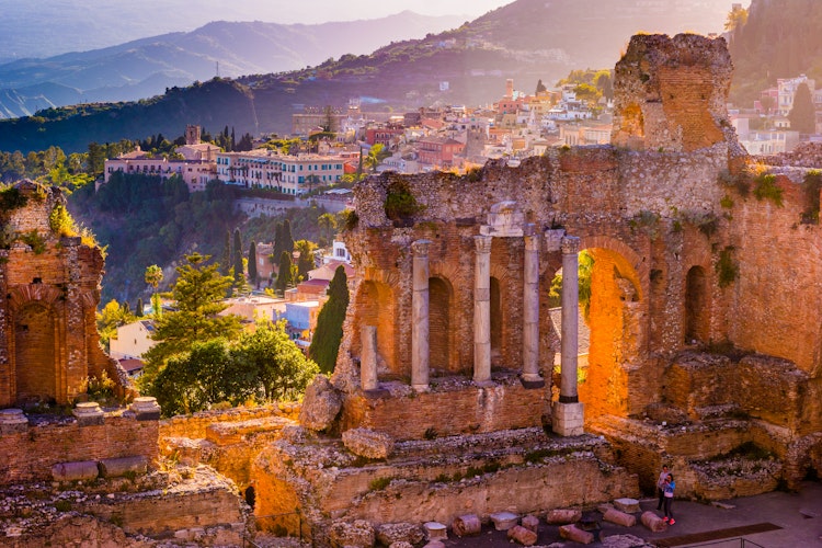 Historiske ruiner af det antikke teater i Taormina badet i gyldent solnedgangslys, med udsigt over Siciliens bjerglandskab og middelhavet i baggrunden