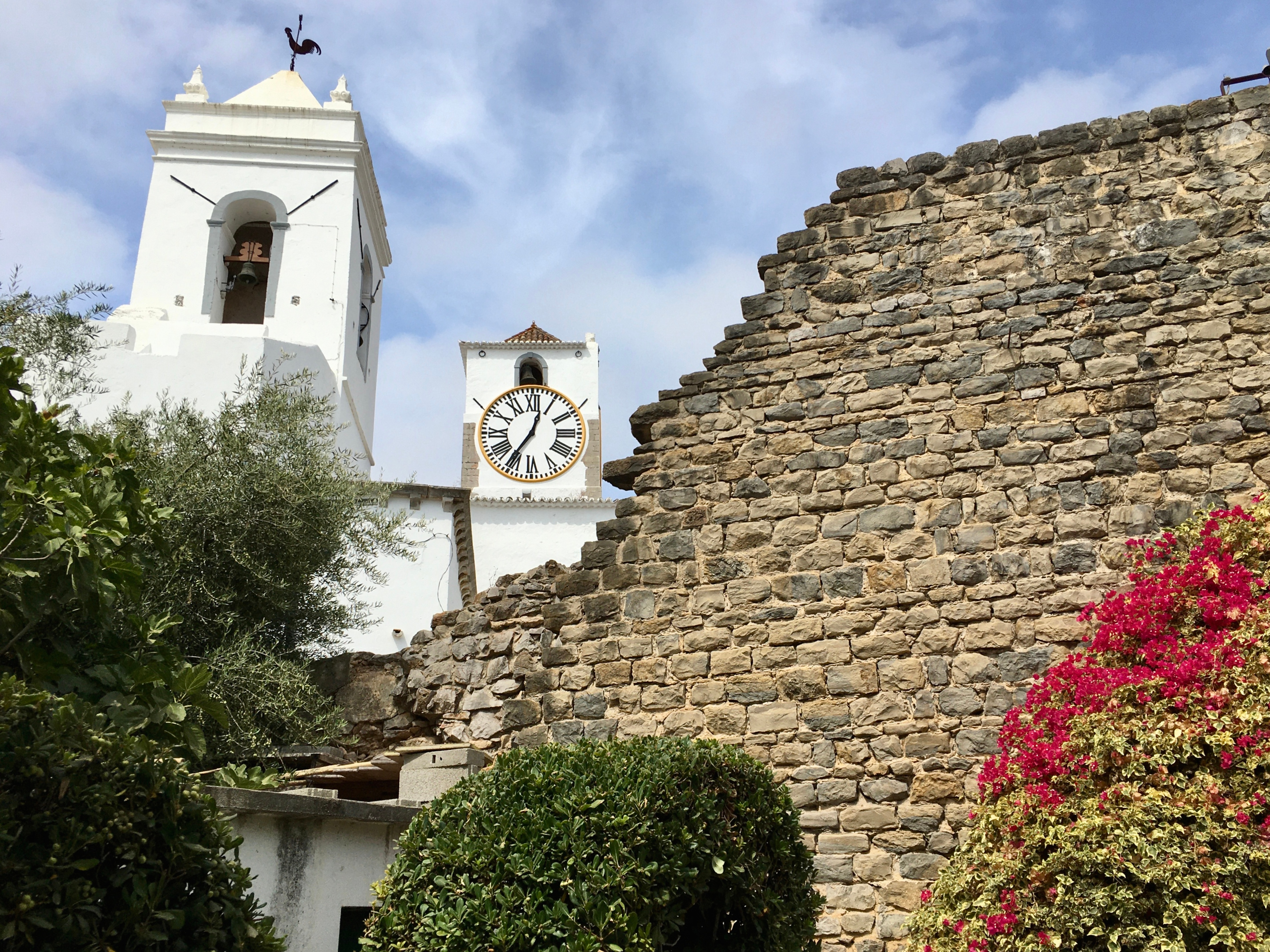 Historisk hvidt kirketårn med ur ved siden af Tavira-slottets gamle stenmure i Algarve, Portugal med lyserøde blomster i forgrunden