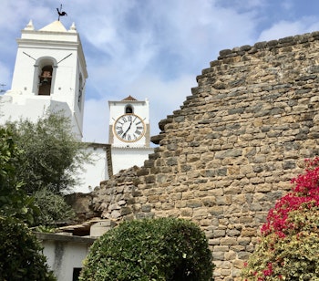 Historisk hvidt kirketårn med ur ved siden af Tavira-slottets gamle stenmure i Algarve, Portugal med lyserøde blomster i forgrunden