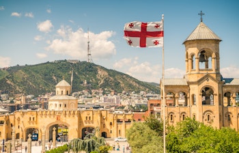 Betagende panoramaudsigt over Tbilisi med det georgiske flag i forgrunden, den historiske Narikala-fæstning på bakketoppen og Kura-floden der snor sig gennem hovedstaden