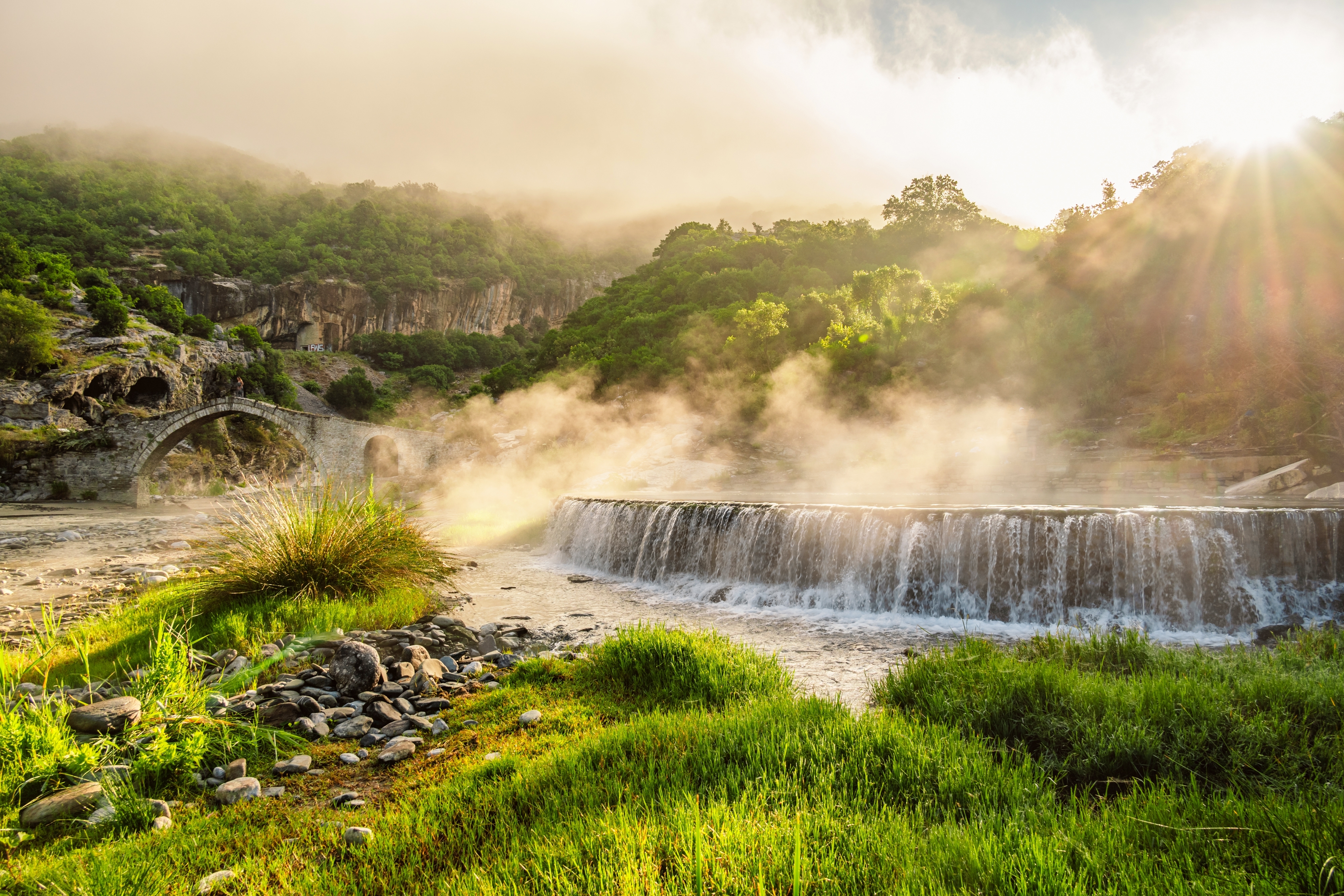 Varmt termisk vandфald i Langarica Canyon nær Permet i Albanien, med den historiske Kadiut Bro synlig gennem tåge og gyldent sollys
