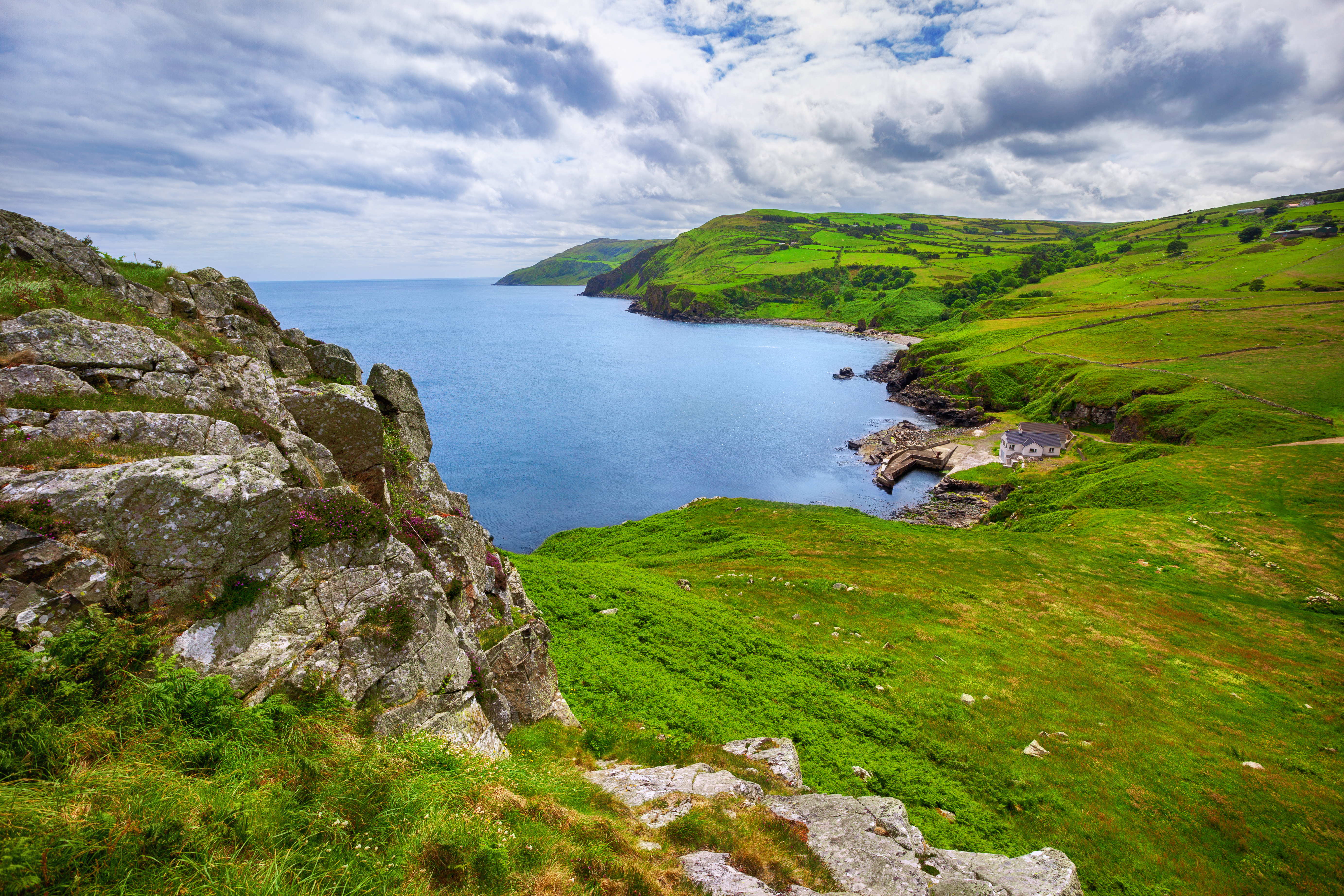 Smuk kystlinje ved Torr Head i County Antrim, Nordirland, med dramatiske grønne klipper, blåt hav og frodige bakkelandskaber under en delvist skyet himmel