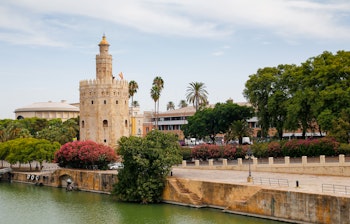 Det historiske Torre del Oro (Guldtårnet) ved bredden af Guadalquivir-floden i Sevilla - et ikonisk andalusisk vartegn fra 1200-tallet omgivet af palmer og frodig vegetation i Spanien