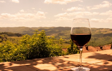 Glas med rubinrød vin i forgrunden med udsigt over Toscanas bølgende bakker dækket af vinmarker i det gyldne solnedgangslys, der indfanger essensen af Italiens vinland