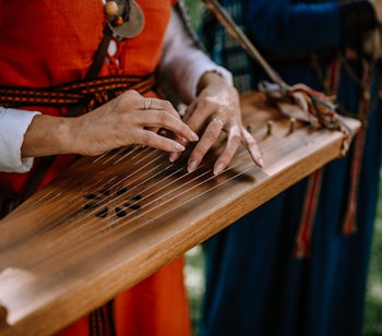 Nærbillede af hænder der spiller på kokle, et traditionelt lettisk strengeinstrument, med musikere i farverige folkekostumer i baggrunden ved en kulturel begivenhed i Valmiera