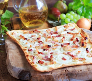 Traditionel tarte flambée fra Alsace med bacon og løg serveret på træbræt med hvidvin, en lokal fransk specialitet