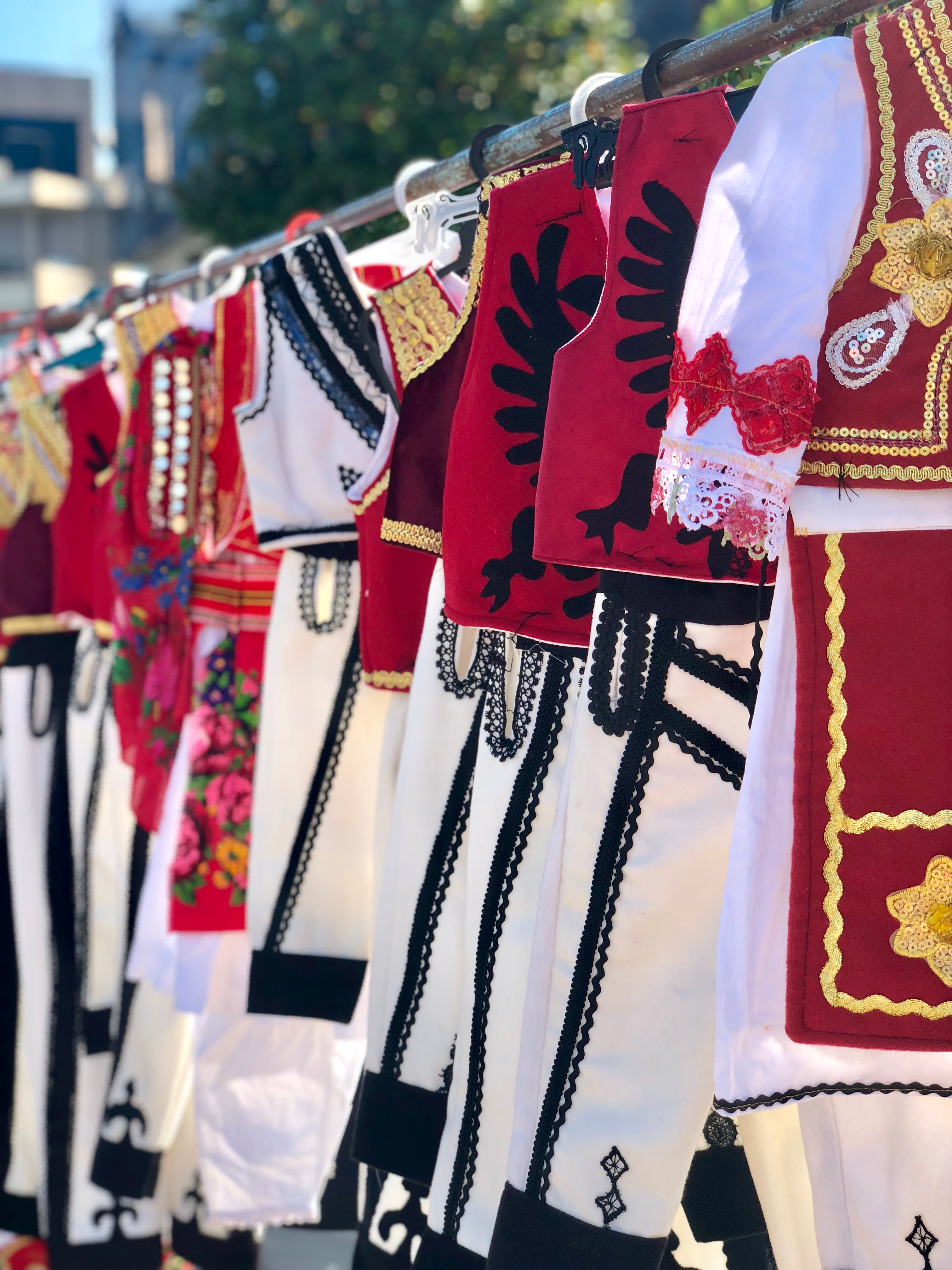 Farverige traditionelle albanske folkedragter med broderede røde veste og hvide klæder udstillet på et gademarked i Shkoder