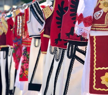 Farverige traditionelle albanske folkedragter med broderede røde veste og hvide klæder udstillet på et gademarked i Shkoder